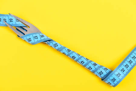 Fork in metallic colour belted tight with turquoise flexible ruler isolated on bright yellow background, copy space. Healthy nutrition conceptの写真素材