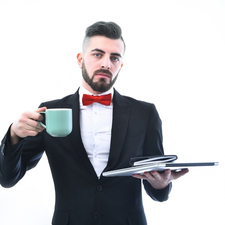 Bearded businessman in suit with concentrated look holds laptop and organizer with light blue cup, isolated on white background. Concept of business confidence and coffee breakの写真素材