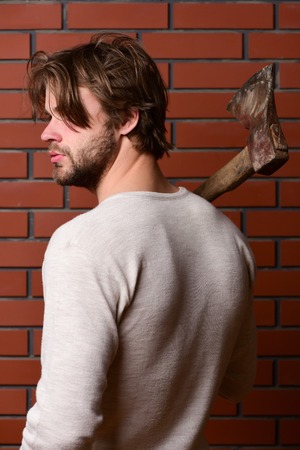 Concept of finished lumbering work with brutal bearded man holding old axe in hand, red brick wall backgroundの写真素材
