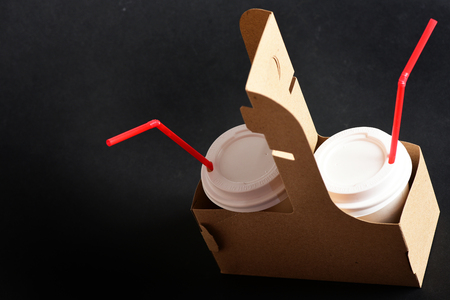 Paper cups with coffee covered with white plastic lids and red straws in eco friendly disposable bag on black background. Takeaway food and drinksの写真素材