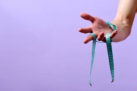 Hand of man holding blue measuring tape tied around its wrist, on purple background, copy space. Fitness, regime and diet concept. Weight management ideaの写真素材