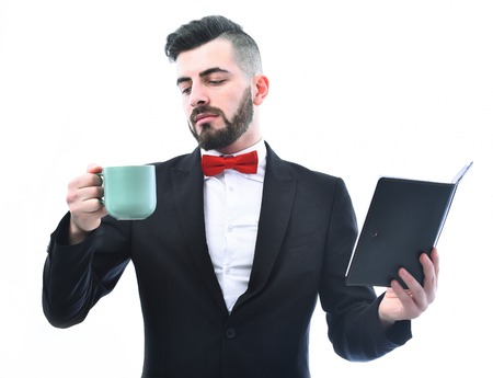 Broker or businessman with beard and smiling face expression holds organizer and looks at cyan coffee cup, isolated on white background. Concept of business breakの写真素材