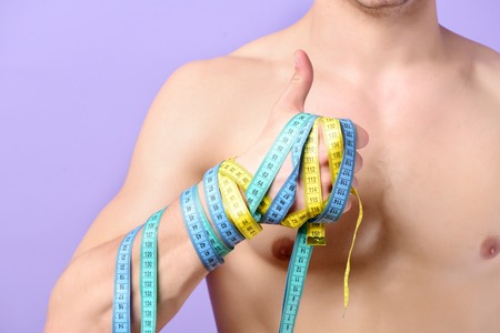 Guys hand with bicolor flexible rulers on light purple background. Mans naked torso and wrist wrapped with cyan and yellow measure tapes showing thumbs up. Fitness, regime and diet conceptの写真素材