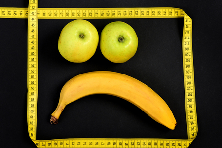 Sad face made of banana fruit and couple of green apples with yellow measuring tape on deep black background. Concept of healthy nutrition, food art and emotionsの写真素材