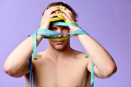 Fitness and diet concept. Weight management idea. Guy with flexible rulers isolated on light purple background. Man with serious face has arms wrapped with yellow and cyan tapes for measuringの写真素材