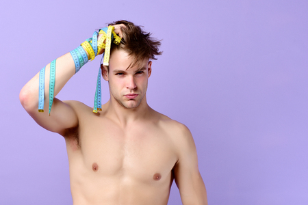 Man with serious and sexy face checks his hair. Guys hand wrapped with yellow and cyan tapes for measuring. Fitness and diet concept. Guy with flexible rulers isolated on purple background, copy spaceの写真素材