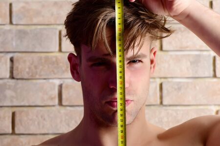Guy with builders ruler near brick wall. Worker with roulette on brick background, defocused. Man with naked torso holds yellow self retracting tape measure. Construction and measurement conceptの写真素材