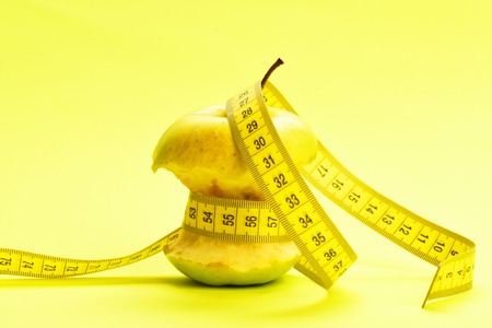 Measured bitten apple. Yellow tape for measuring wraps around bitten apple on light yellow backgroundの写真素材