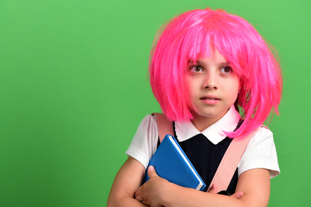 School girl in pink wig with surprised face on green background. Girl holds blue book. Back to school and education concept. Pupil in school uniform with pink backpack, copy spaceの写真素材