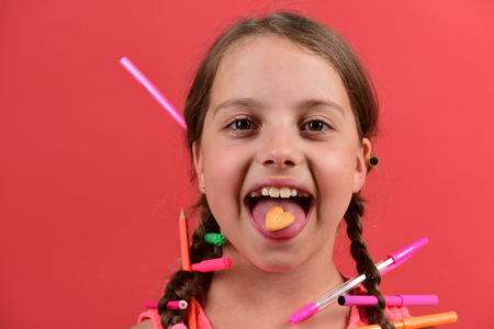 School girl with happy face isolated on salmon red background. Back to school concept. Kid in pink dress with braids and colored pencils in them. Girl holds heart shaped cookie on tongueの写真素材
