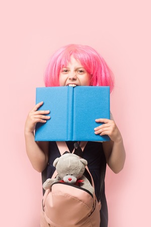 Girl with backpack and toy bites big blue book. Schoolgirl with pink wig isolated on light pink background. Kid with angry face in school uniform. Childhood and back to school concept.の写真素材