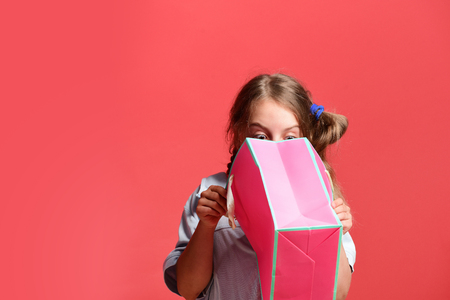 School girl with package isolated on salmon red background. Sale and shopping concept. Girl with pink shopping bag looks inside it. Kid with surprised sight and stylish hairdo does shoppingの写真素材