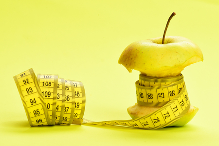 Concept of weight management. Bitten apple wrapped around with measuring tape on light yellow background as symbol of tasty and dietary foodの写真素材