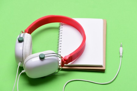 Headphones in white and red color with empty notebook. Music accessories and note taking concept. Modern and stylish earphones isolated on light green background. Headset for music and blank pageの写真素材