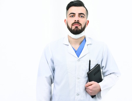 Doctor with beard holds notebook with history of present illness. Treatment and ambulance services concept. Man with confident face in white medical coat. Surgeon in mask isolated on white backgroundの写真素材