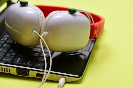 Headphones and black laptop. Electronics isolated on light yellow background. Sound recording idea. Earphones in red and white colors made of plastic with computer. Music and digital equipment conceptの写真素材