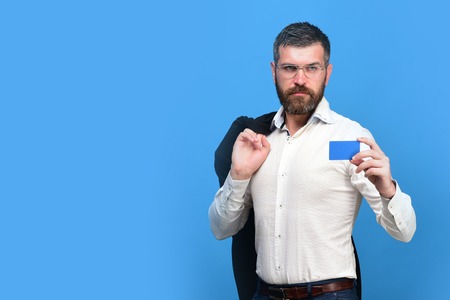 Man with beard in white shirt holds dark blue business card. Business and people at work concept. Guy with serious face and glasses isolated on blue background. Businessman with empty card, copy spaceの写真素材
