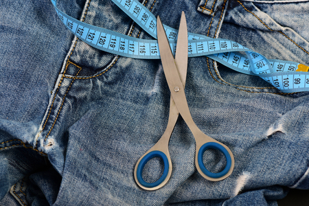 Tailors tools on denim textile. Making clothes and design concept. Jeans belt loops, zipper and pocket, close up, selective focus. Metal scissors and blue measure tape on jeans.の写真素材