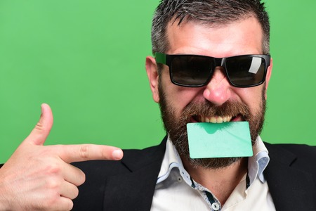 Man with dark beard holds green business card in teeth. Guy with funny face and sunglasses isolated on green background. Business and success concept. Businessman points at empty card, copy spaceの写真素材