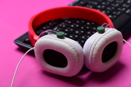 Earphones in red and white colors with computer keyboard. Sound recording idea. Electronic appliances on pink background, selective focus. Music and call center concept. Headphones and black keyboardの写真素材