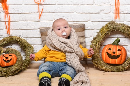 small baby boy with adorable curious face in yellow sweater leaned on pillow in knitted scarf around halloween decorations on white brick wall backgroundの写真素材