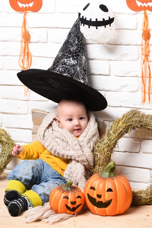 small baby boy with adorable curious face in yellow sweater leaned on pillow in knitted scarf and black witch hat around halloween decorations on white brick wall backgroundの写真素材