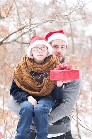 small boy in glasses, santa claus hat and knitted scarf and handsome man or bearded father guy holds red christmas or new year present box in winter outdoor with snow on natural backgroundの写真素材