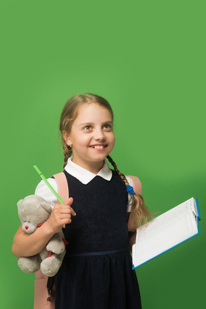 Girl with braids and happy face. Pupil holds blue book, marker and teddy bear. Kid in school uniform isolated on green background. Study and back to school concept.の写真素材