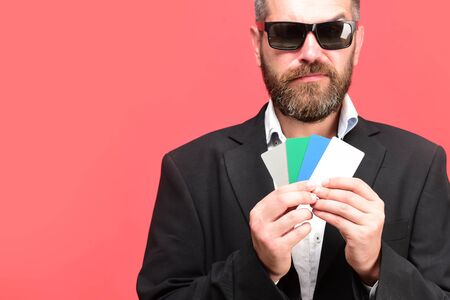 Business and luck concept. Guy with tricky face and sunglasses isolated on pink background. Man with beard holds colorful business card. Businessman with empty cards, copy spaceの写真素材