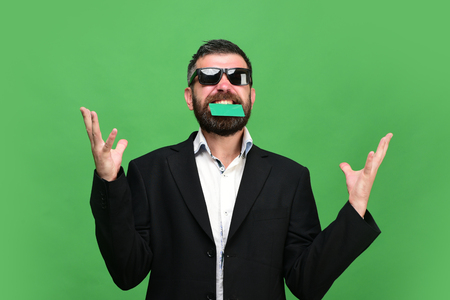 Man with dark beard in suit holds green business card in teeth. Guy with happy face and sunglasses isolated on green background. Businessman with empty card. Business and success conceptの写真素材