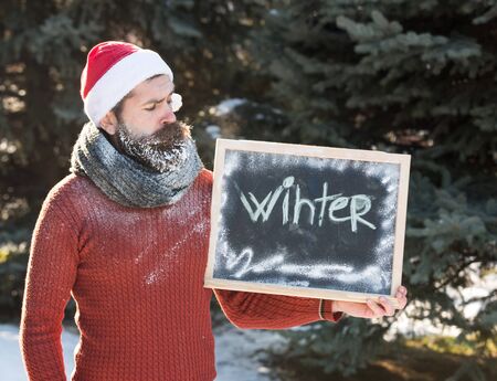man in santa claus hat, bearded hipster with beard and moustache covered with white frost, gives ok gesture with winter word on black board or chalkboard on sunny day on natural backgroundの写真素材