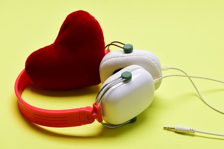 Headset for music with love symbol. Headphones in white and red color and soft toy heart. Modern and stylish earphones on light yellow background. Love, leisure and music conceptの写真素材