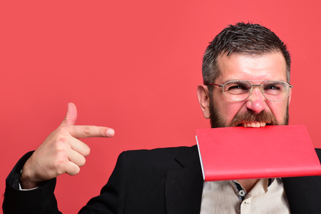 Success, business and scheduling concept. Businessman with funny face and glasses isolated on red background. Guy bites closed notebook and points at it. Man with beard holds red organizer in teethの写真素材