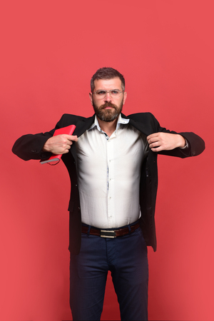 Businessman with strict face and glasses on light red background. Guy with closed red notebook. Success, business and studying concept. Man in classic outfit with beard holds red book or organizerの写真素材