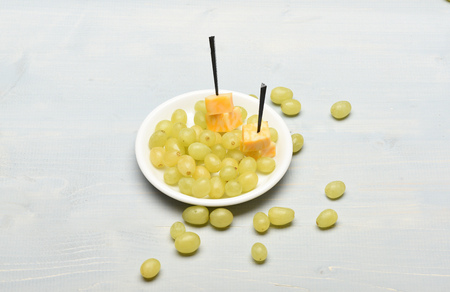 Gourmet food concept. White grapes on plate and around it. Delicious appetizer with cheese and summer fruit, close up. Composition of marble cheese skewers and grapes on vintage background.の写真素材