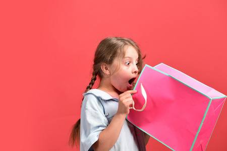 Fashion and shopaholism concept. Girl holds pink shopping bag and looks inside. Kid with surprised face and stylish hairdo does shopping. School girl with package isolated on salmon red backgroundの写真素材