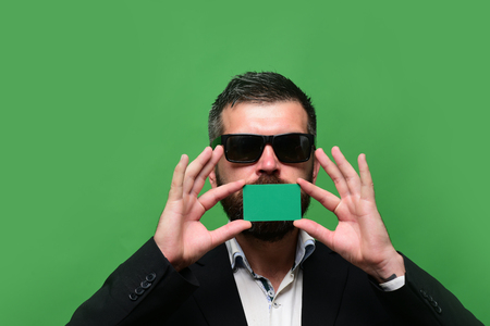 Guy with serious face and sunglasses isolated on green background. Businessman with empty card, copy space. Man with beard holds green business card near mouth. Business and success conceptの写真素材