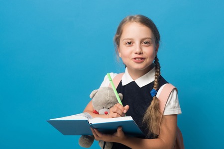Girl with braids and happy face. Kid in school uniform isolated on blue background with copy space. Pupil writes in blue book with green marker and holds teddy bear. Study and back to school concept.の写真素材