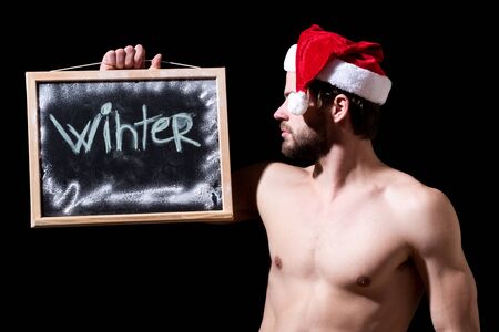 handsome bearded man or sexy christmas guy with muscular body and chest in red santa claus hat holds blackboard or chalkboard with winter text on black background, copy spaceの写真素材