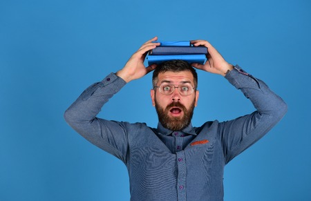 Knowledge and scheduling concept. Man with beard and books. Teacher wears glasses and holds notebooks. Textbooks in blue color on guys head isolated on blue background. Professor with surprised faceの写真素材