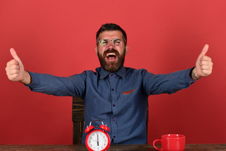Deadline and great job concept. Man with beard and glasses shows thumbs up, red background. Worker with happy smiling face sits at wooden table. Tea cup and retro clock on vintage tableの写真素材