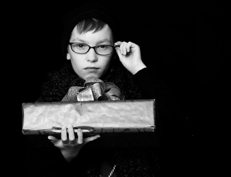 small boy or cute nerd kid in glasses, hat and fashionable knitted scarf on black background holds red present box with golden bowの写真素材