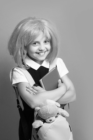 Girl with backpack, book and toy. Schoolgirl with pink wig isolated on green background. Childhood and back to school concept. Kid with happy face in school uniform.の写真素材