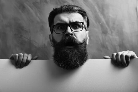 Bearded man, long beard. Brutal caucasian unshaven surprised hipster with glasses and moustache holding yellow paper sheet on brown studio backgroundの写真素材