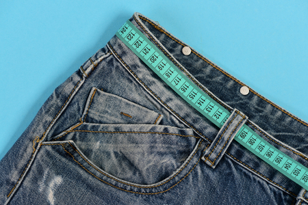 Healthy lifestyle and dieting concept. Top part of denim trousers isolated on blue background. Jeans belt loops and pocket in close up. Jeans with blue measure tape instead of belt.の写真素材