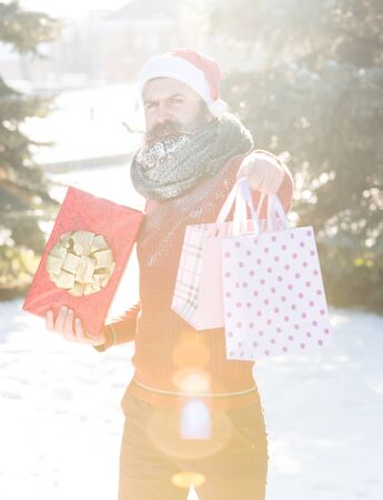 Handsome man in santa claus hat, bearded hipster with beard and moustache covered with white frost, holds red gift box and bags as presents on bright sunny winter day outdoors on natural backgroundの写真素材