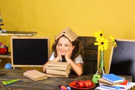 Girl sits at desk with books, flowers, fruit and colorful stationery. Kid and school supplies on yellow background. Schoolgirl with happy face and book on her head. Childhood and study time conceptの写真素材