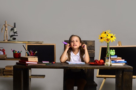 Kid and school supplies on grey wall background. Childhood and study time concept. Girl sits at her desk with books, flowers and colorful stationery. Schoolgirl with thinking face draws in art bookの写真素材