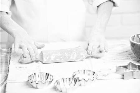 young male hands of cook chef in white uniform standing near table with rolling pin molds for cakes red bowl and lot of flour and cooking on brick wall backgroundの写真素材