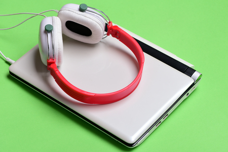 Sound recording idea. Electronics isolated on light green background. Earphones in red and white colors made of plastic with computer. Music and digital equipment concept. Headphones and silver laptopの写真素材